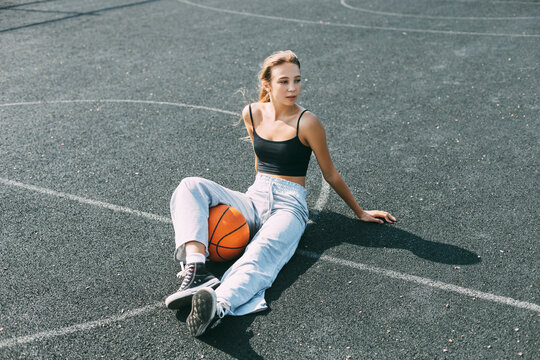 A basketball player is sitting on a sports field with a basketball and looks away. Sports, fitness, lifestyle