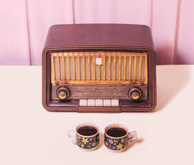 Nostalgia concept made of antique radio from 50s and two retro cups filled with fresh hot blend of black coffee. Arrangement made against baby pink and beige background.