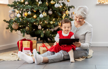 Obraz na płótnie Canvas winter holidays and family concept - happy grandmother and baby granddaughter with tablet pc computer sitting near christmas tree at home