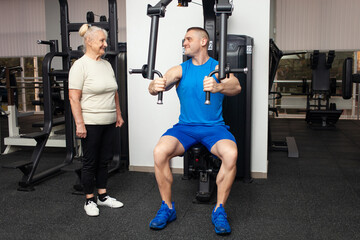 Fototapeta na wymiar handsome young coach man shows senior woman how to properly perform exercises on the simulator in the gym. Happy people smile, enjoy, happy. Active sports healthy lifestyle indoor. Sports uniform