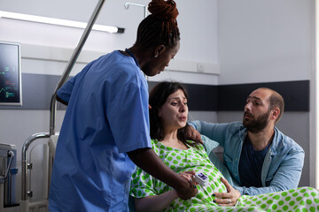 Pregnant woman in pain getting medical assistance from black nurse while sitting in hospital ward bed. Young man supporting wife preparing for childbirth. Multi ethnic people with child delivery