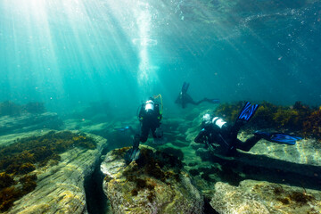 Fototapeta na wymiar Scuba Divers Kick Over Rocks Underwater in Sun Light with Bubbles