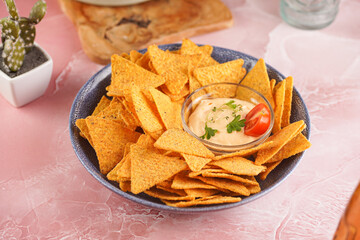 A blue bowl with cheese tortilla chips and a chilli cheese dip on a pink marble surface, cherry tomatoes, ketchup