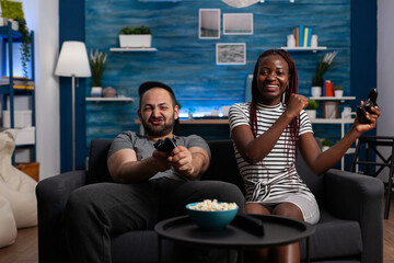 Cheerful interracial people winning video game on TV while playing with console and joysticks. Mixed race couple using technology for entertainment, having fun on sofa using controllers