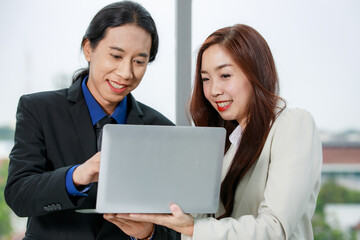 Asian young happy successful long ponytail hair businessman and confident businesswoman colleague stand smiling holding laptop computer together planning discuss about work online in company office