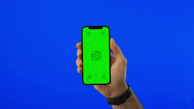 Hand with the Phone Appears from the Bottom. Man Holding a Phone with Vertical Green Chroma Key Screen Isolated on Blue Background.