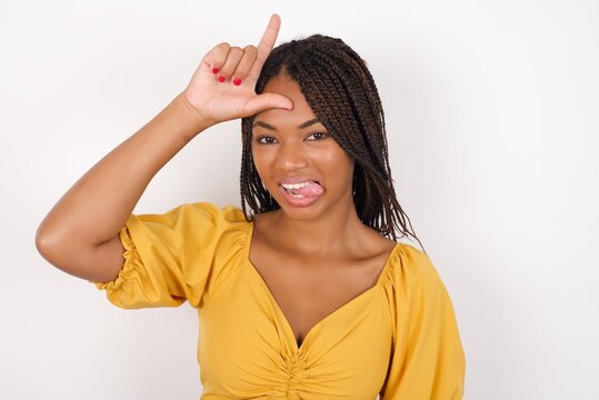 Funny Young african american woman with braids over white wall makes loser gesture mocking at someone sticks out tongue making grimace face.