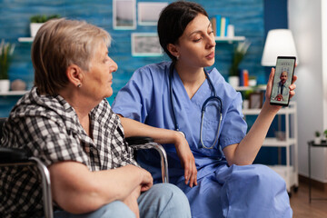 Nursing home female worker helping retired disabled elderly woman to have a video call on smartphone with her doctor. Social assistant working with people with disability in retirement home