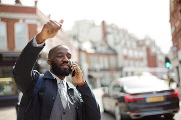 Young businessman talking on cell phone and hailing taxi on sunny urban street