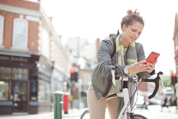 Young woman commuting on bicycle, texting with cell phone on sunny urban street