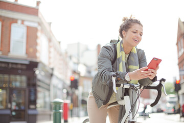 Young woman commuting on bicycle, texting with cell phone on sunny urban street