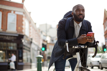 Young businessman commuting with bicycle, texting with cell phone on sunny urban street