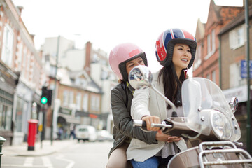 Smiling young women friends wearing helmets, riding motor scooter on urban street