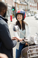 Smiling young woman in helmet on motor scooter, talking to friend on urban street