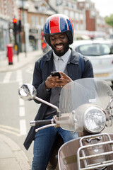 Smiling young businessman in helmet on motor scooter texting with cell phone on urban street