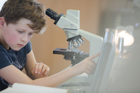 Boy student conducting scientific experiment at microscope and computer in laboratory classroom