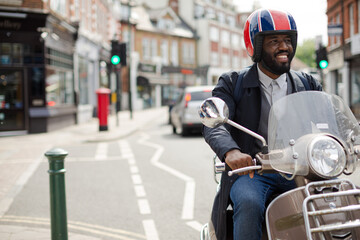 Smiling young businessman in helmet riding motor scooter on urban street