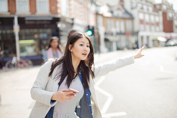 Fototapeta premium Young businesswoman with cell phone hailing taxi on sunny urban street