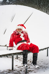 Christmas holiday and active lifestyle. Funny Santa Claus in red traditional costume sits on a trampoline in a winter snowy amusement park.