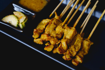 Pork satay with side dish and peanut dipping sauce in a black plate isolated on black background.