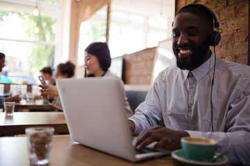 Man with headphones using laptop and drinking coffee in cafe
