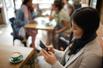 Young woman with headphones texting with cell phone and drinking coffee at cafe table