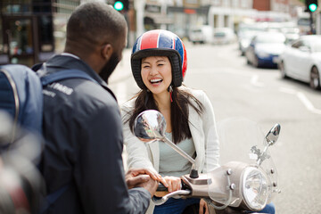 Smiling young woman in helmet on motor scooter, talking to friend on urban street
