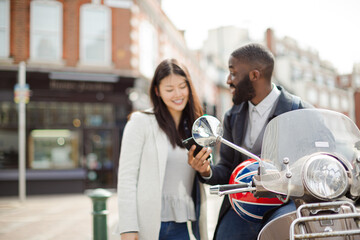 Young couple laughing, using cell phone at motor scooter on sunny urban street