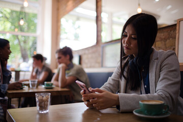 Young woman with headphones texting with cell phone and drinking coffee at cafe table