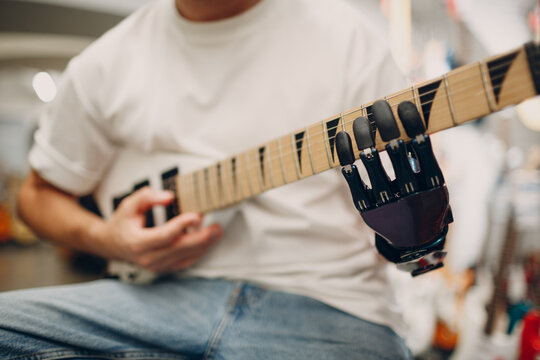 Young disabled man with artificial prosthetic hand plays on electric guitar at musical instruments shop