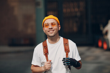 Young disabled man with artificial prosthetic hand in casual clothes and backpack walking at sunny...