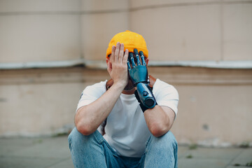 Young disabled upset sad man with artificial prosthetic hand close his face sitting on sidewalk...