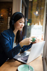 Pensive young woman at laptop listening to music with headphones at cafe window