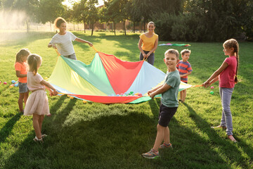 Group of children and teachers playing with rainbow playground parachute on green grass. Summer...
