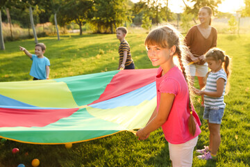 Group of children and teacher playing with rainbow playground parachute on green grass. Summer camp activity - obrazy, fototapety, plakaty
