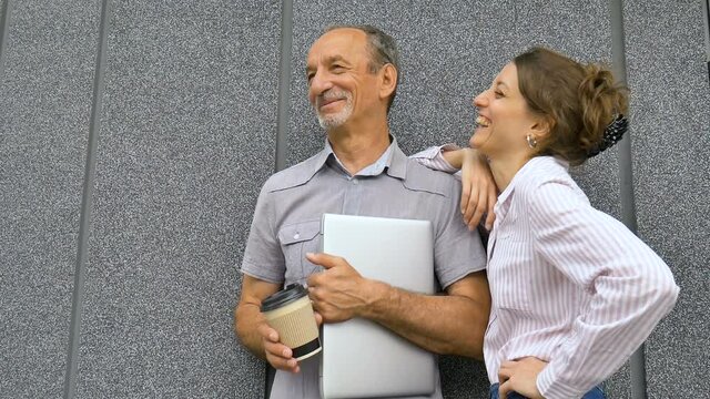 Young woman and senior man are waiting for a meeting standing near the grey wall outdoors in the morning. Colaboration between older coworker and younger colleage