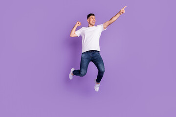 Full length photo of happy young cheerful man jump up point finger empty space isolated on purple color background