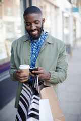 Young man with coffee and shopping bags texting with cell phone on urban sidewalk