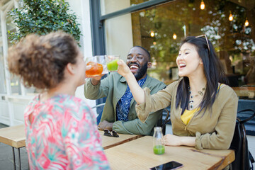 Smiling enthusiastic friends toasting fresh juice glasses at sidewalk cafe