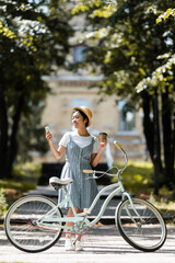 Obraz premium smiling asian woman with takeaway drink using smartphone near bike in park