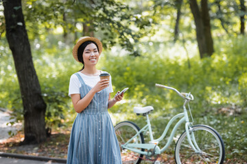 happy asian woman with takeaway drink and mobile phone looking at camera near bike in park