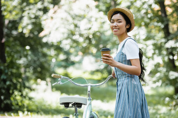 happy asian woman with takeaway drink looking at camera near bicycle in park