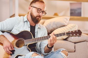 Sitting near the sofa. Man in casual clothes and with acoustic guitar is indoors