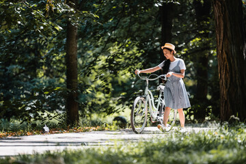 Obraz premium happy asian woman in striped sundress walking with bicycle in park