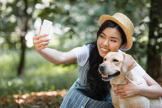joyful asian woman in straw hat taking selfie with yellow labrador in park
