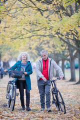 Senior couple walking bicycles among trees and leaves in autumn park