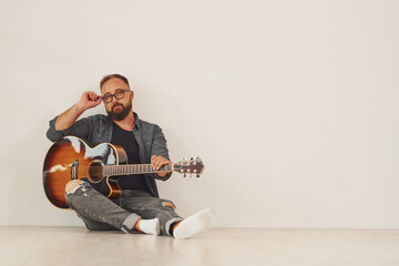 Obraz premium Sitting against wall. Man in casual clothes and with acoustic guitar is indoors
