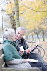 Senior couple reading newspaper and drinking coffee on bench in autumn park