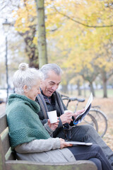 Senior couple reading newspaper and drinking coffee on bench in autumn park