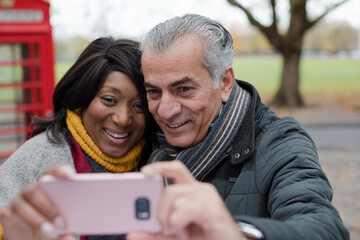 Smiling senior couple taking selfie in autumn park in front of red telephone booths